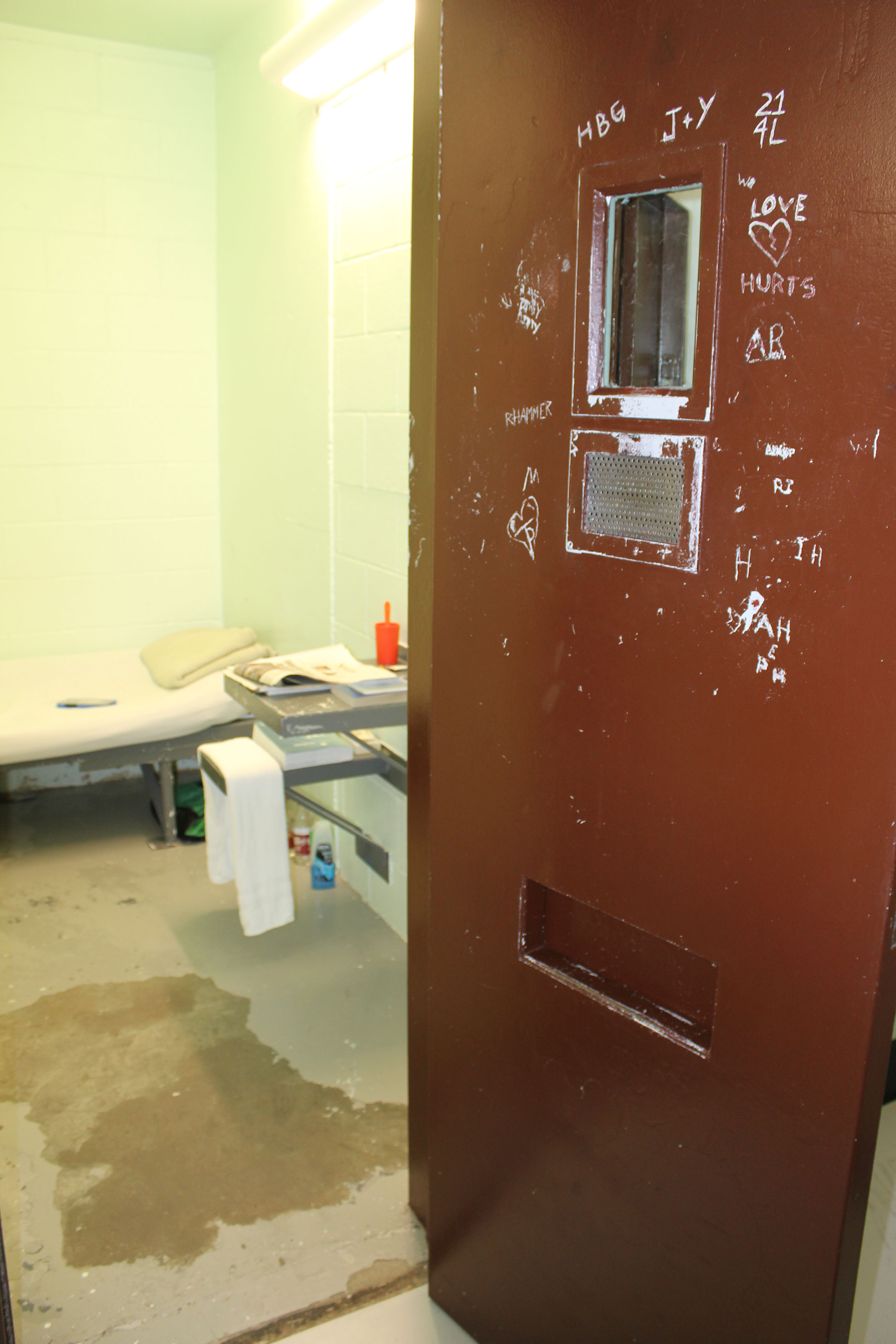 A metal door that has been painted dark brown has initials scratched into it. Behind the door you can see some of a small prison cell.