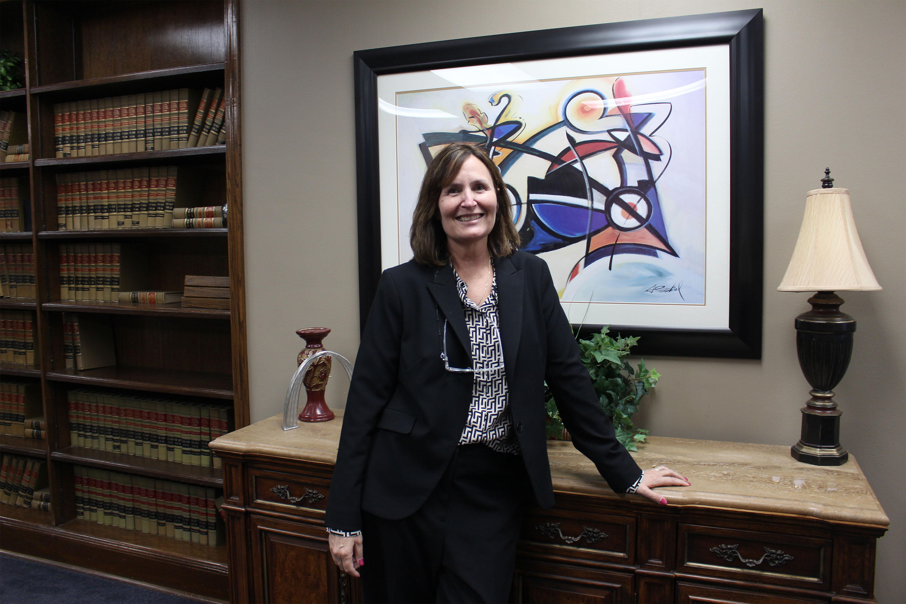 A woman in a dark suit stands with her left hand resting on a chest of drawers and smiles at the camera. There is a painting on the wall behind her and a bookshelf with law books.