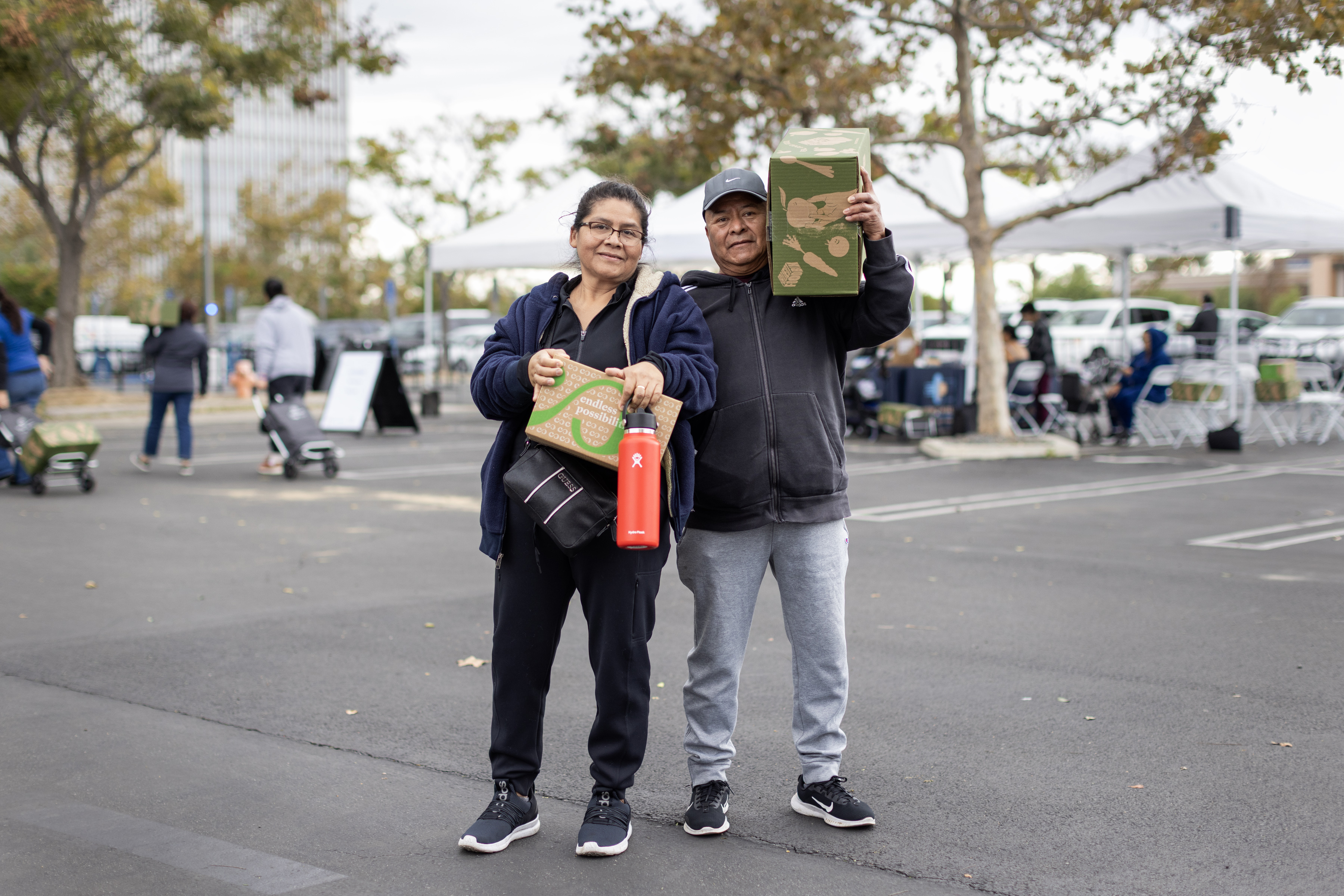 A Hispanic couple stands outside. The woman on the left holds a cardboard box and water bottle. Her husband stands to the right of her, carrying another box on his shoulder.